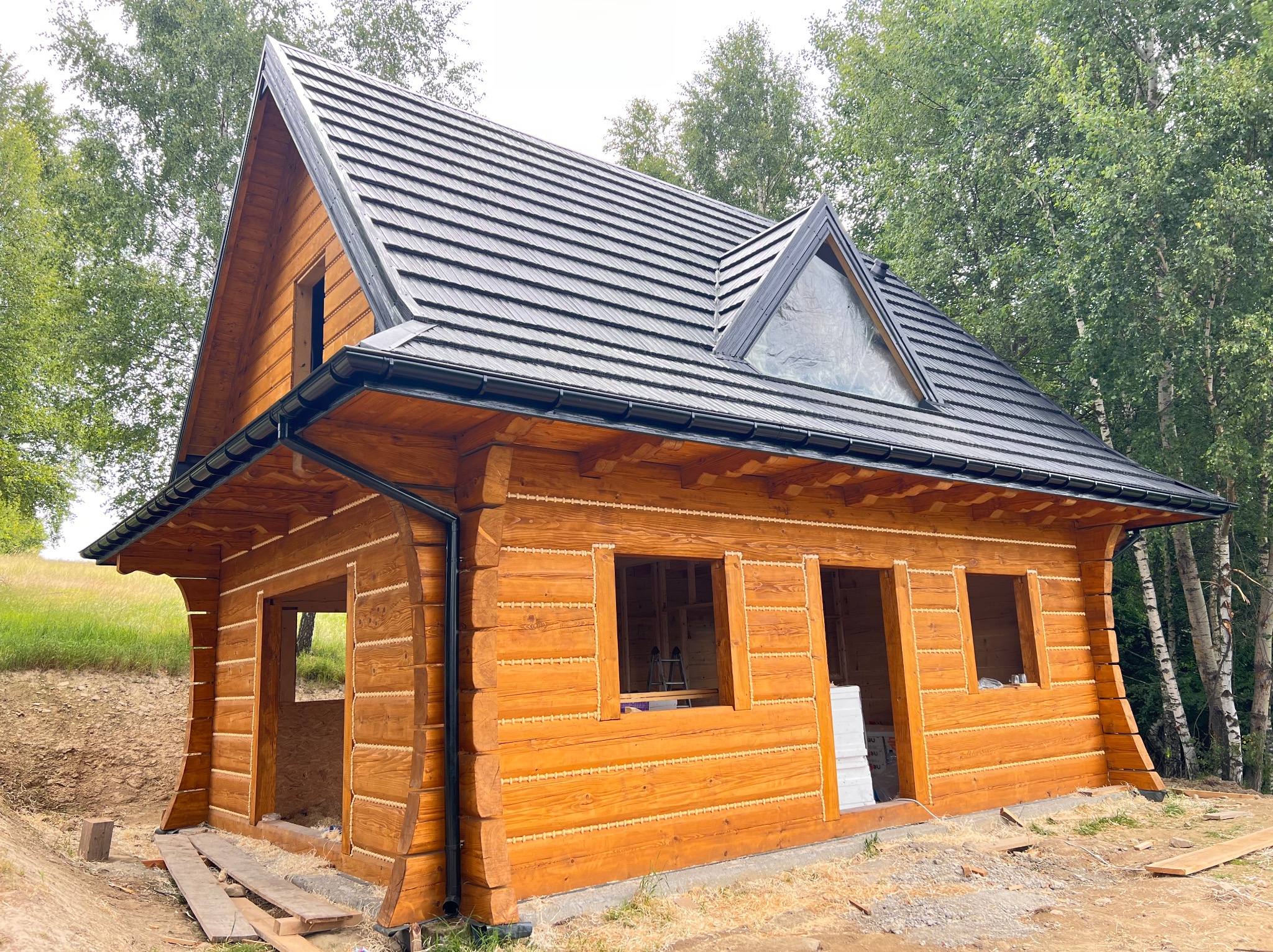 Surowa bryła domu z bali, z ciemnym dachem i oknem trójkątnym na poddaszu, otoczona zielenią. Widoczne detale konstrukcyjne i brakujące elementy wykończenia.