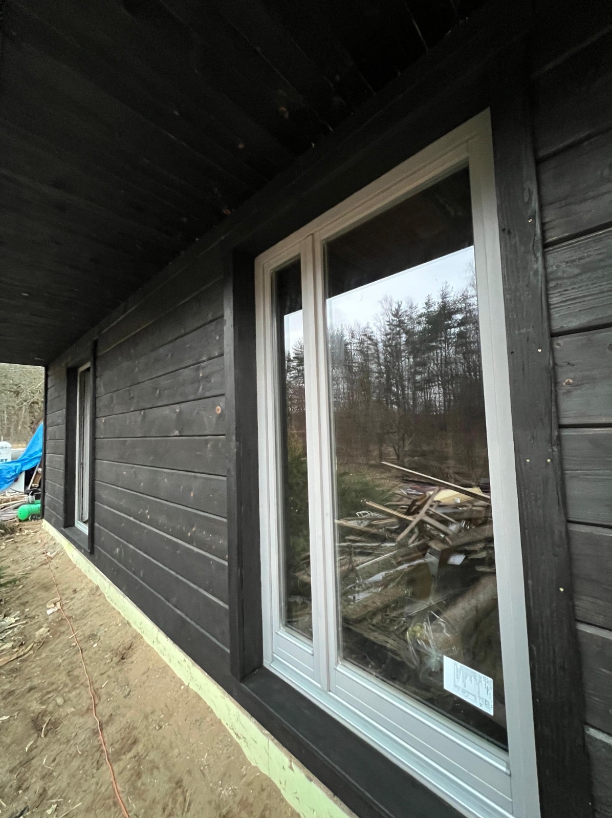 Ciemna, drewniana elewacja domu z białymi oknami. Widoczne deski elewacyjne i fragment terenu budowy. W oknach odbija się las.