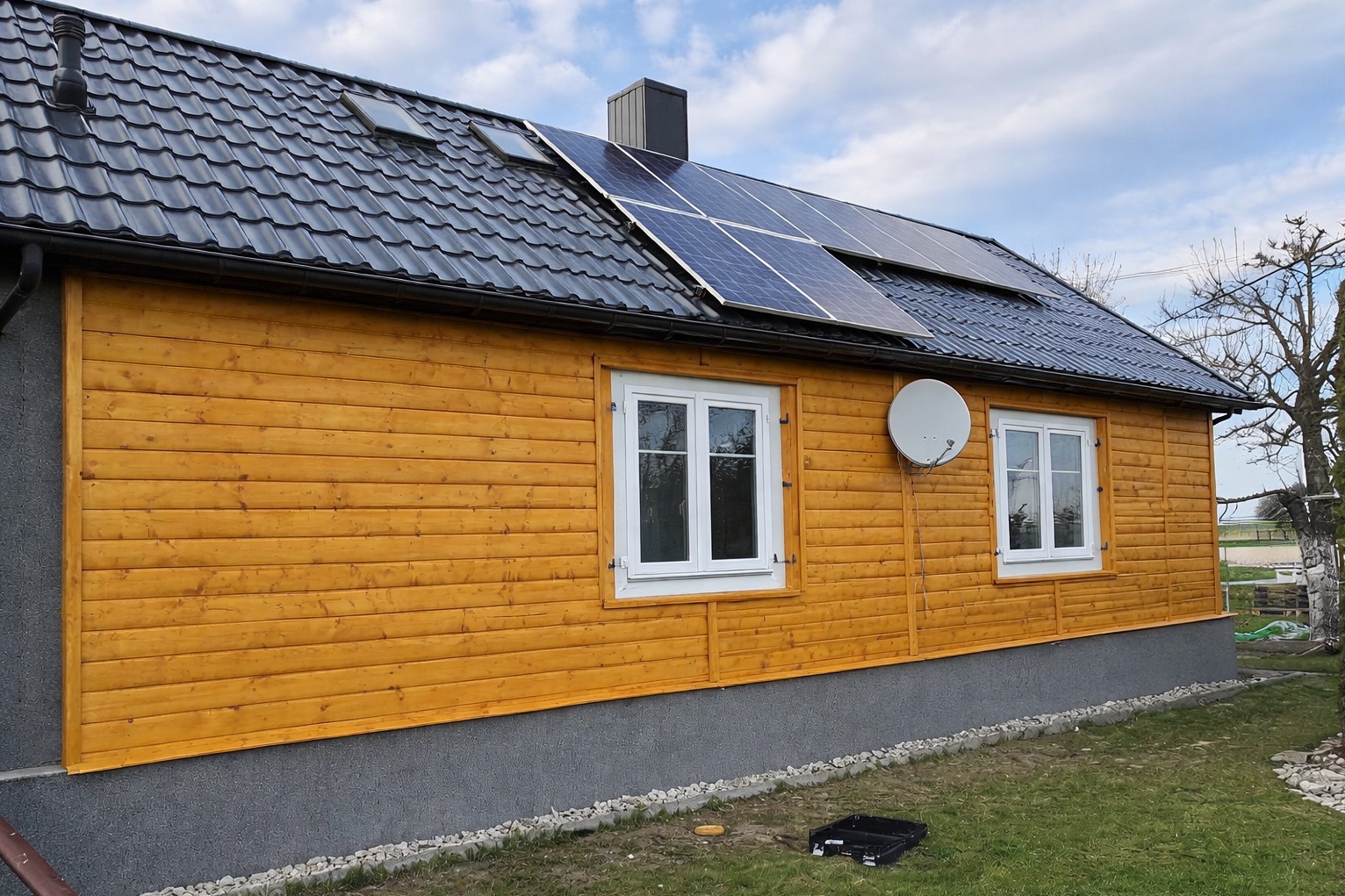 Dom z drewnianą elewacją w odcieniach brązu, czarnym dachem z panelami słonecznymi i białymi oknami. Widoczna antena satelitarna i fragment ogrodu.