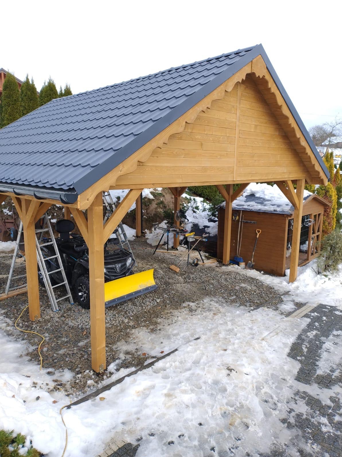 Drewniana wiata garażowa z szarym dachem, częściowo ośnieżona, z pojazdem ATV z żółtym pługiem w środku. Obok widoczny mały drewniany budynek gospodarczy.
