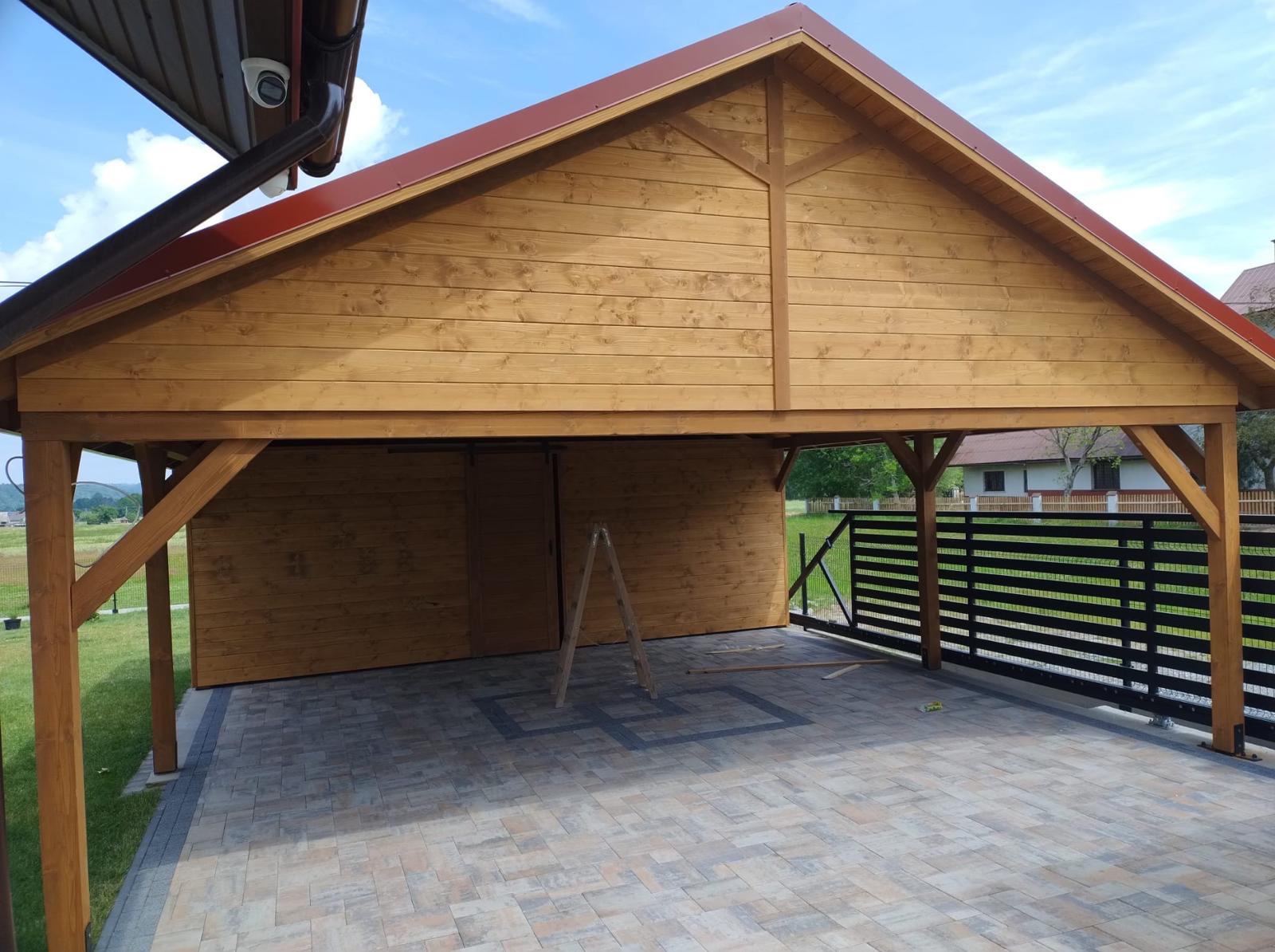 Drewniana konstrukcja z zadaszeniem i przyległym ogrodzeniem panelowym na kostce brukowej. Widoczna drabina oparta o ścianę. Ujęcie w słoneczny dzień.