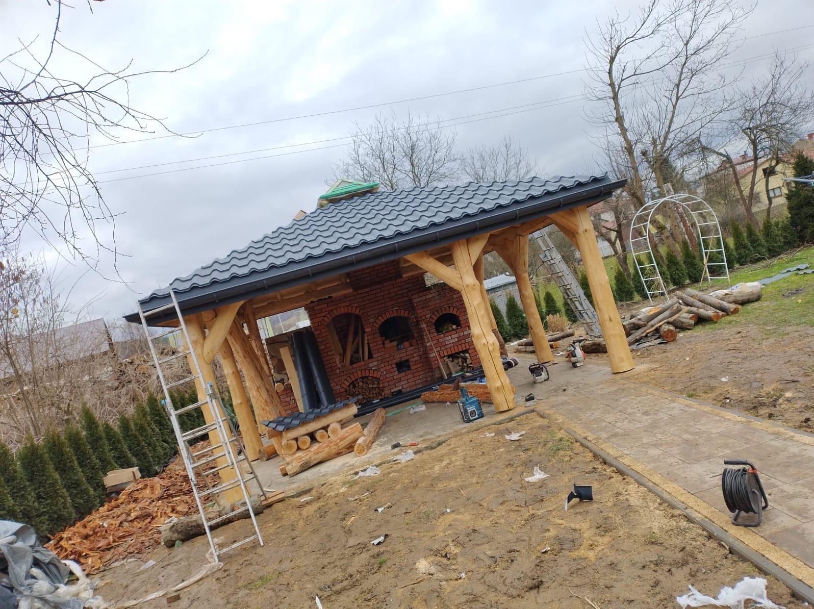 Otwarta altana z ceglanym grillem i drewnianymi słupami, pokryta szarą dachówką. Widoczne narzędzia i drabina sugerują etap końcowy budowy.