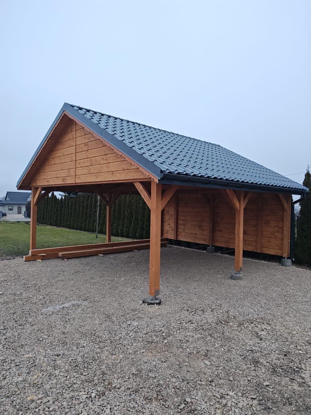 Drewniana wiata z szarym dachem na żwirowym podłożu. Widoczne elementy konstrukcyjne i fragmenty niezabudowane. W tle zieleń i budynki.