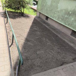Tilawi - Teren z wyrównaną, świeżo spulchnioną ziemią pod nasadzenia, wzdłuż chodnika z zieloną barierką i fragmentem jasnej elewacji.