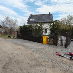 BS Garden - Dom z ciemnym dachem, otoczony zielenią i ogrodzeniem. Na pierwszym planie narzędzia ogrodnicze i żwirowa droga. Błękitne niebo z chmurami w tle.