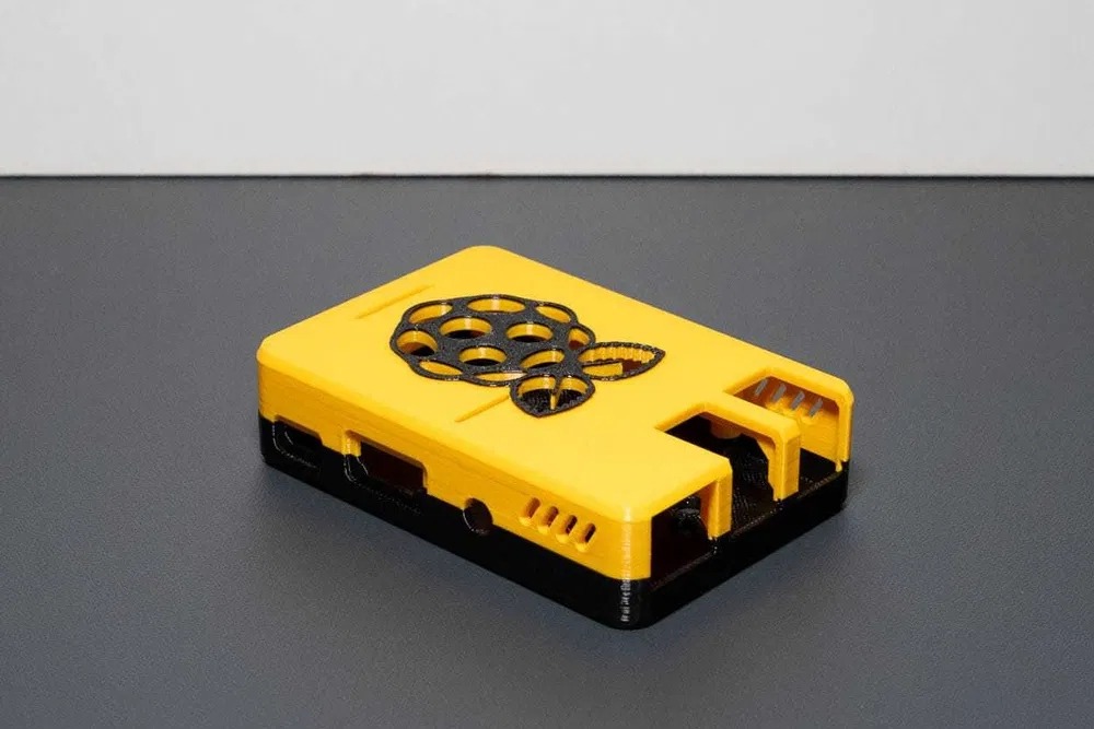 Żółto-czarna obudowa Raspberry Pi z logo maliny, wykonana metodą druku 3D. Widoczne otwory wentylacyjne i porty. Precyzyjne wykonanie, gładka powierzchnia.