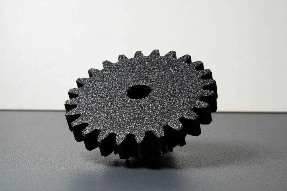 Czarny, teksturowany model koła zębatego z otworem pośrodku, wydrukowany przestrzennie, leżący na szarym tle z białym tłem w tle. Widoczne detale powierzchni.