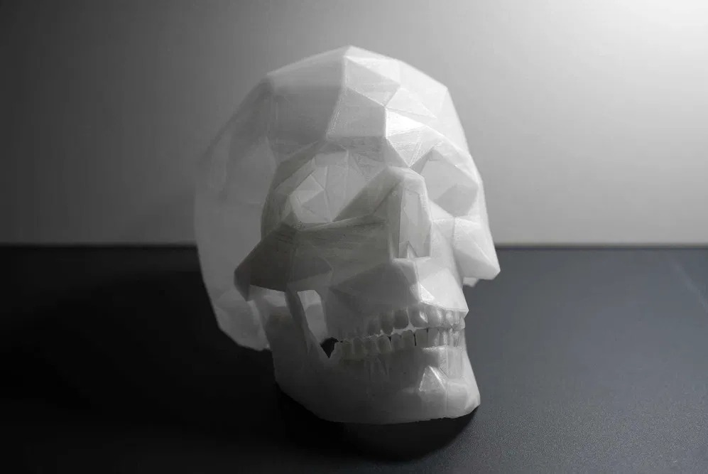 Biała, geometryczna czaszka, wydrukowana w 3D, ukazana z boku na ciemnym tle. Widoczne fasetowe powierzchnie i detale zębów. Monochromatyczna kolorystyka.