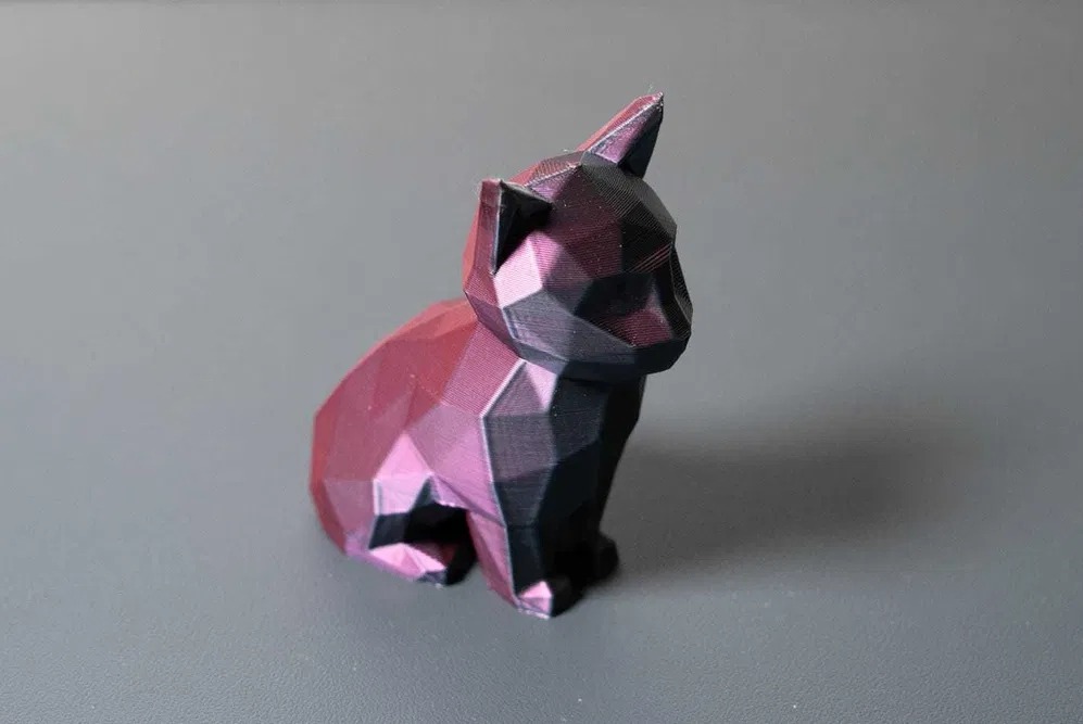 Figurka kota wydrukowana w 3D, w geometrycznym stylu, z widoczną strukturą druku, w kolorach różu i czerni, na szarym tle.