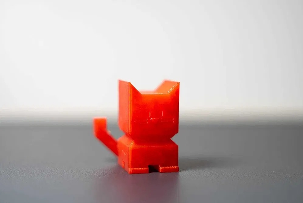 Czerwona, geometryczna figurka kota, wydrukowana w 3D, na szarym tle. Widoczne warstwy druku, ostre krawędzie i uproszczony design.