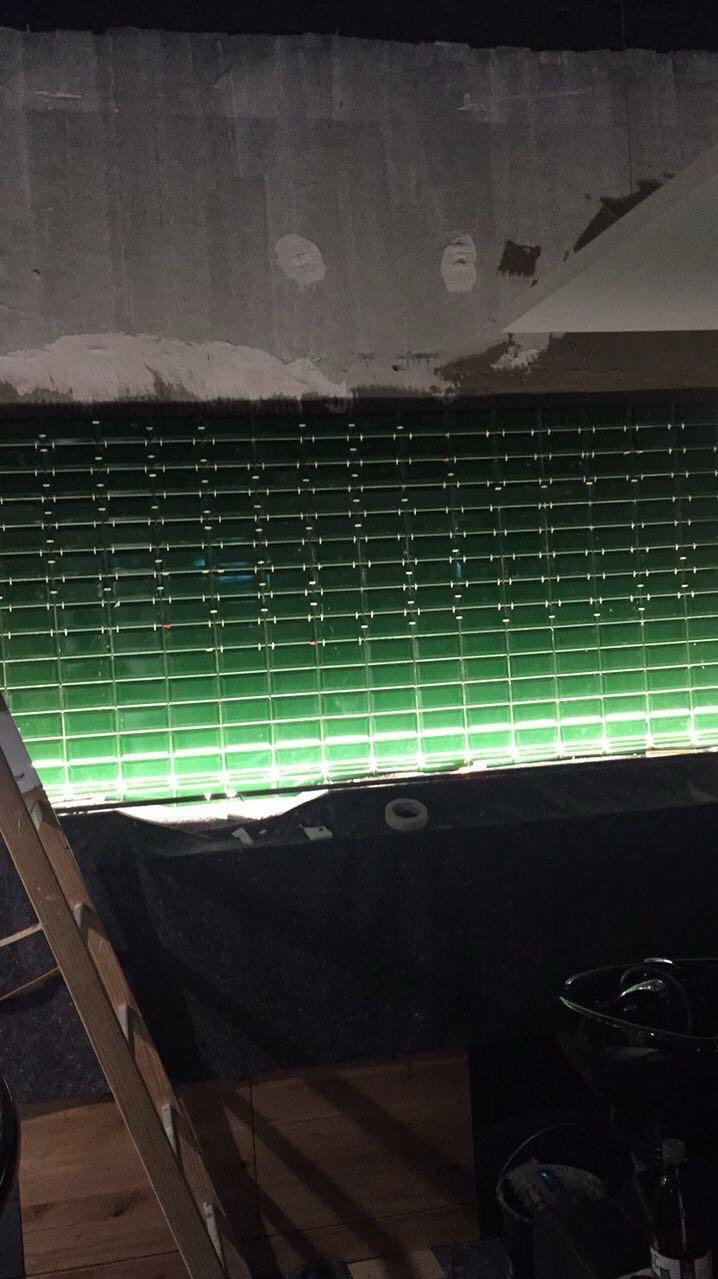 Ściana wyłożona błyszczącą, zieloną mozaiką w trakcie prac. Widoczne nierówności sufitu i drabina oparta o ścianę. Oświetlenie podkreśla fakturę płytek.