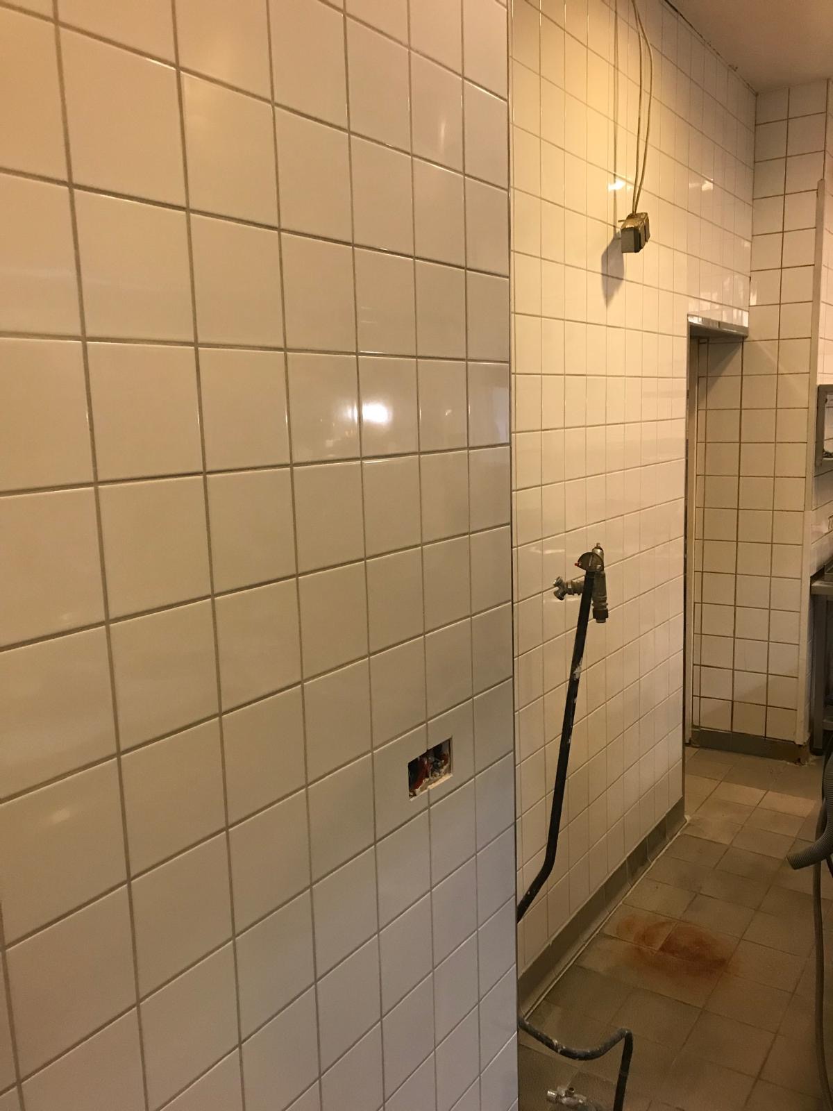 Wykończona ściana z białych płytek w kuchni, widoczne przyłącze wodne i otwór na gniazdko elektryczne. Podłoga wyłożona płytkami.