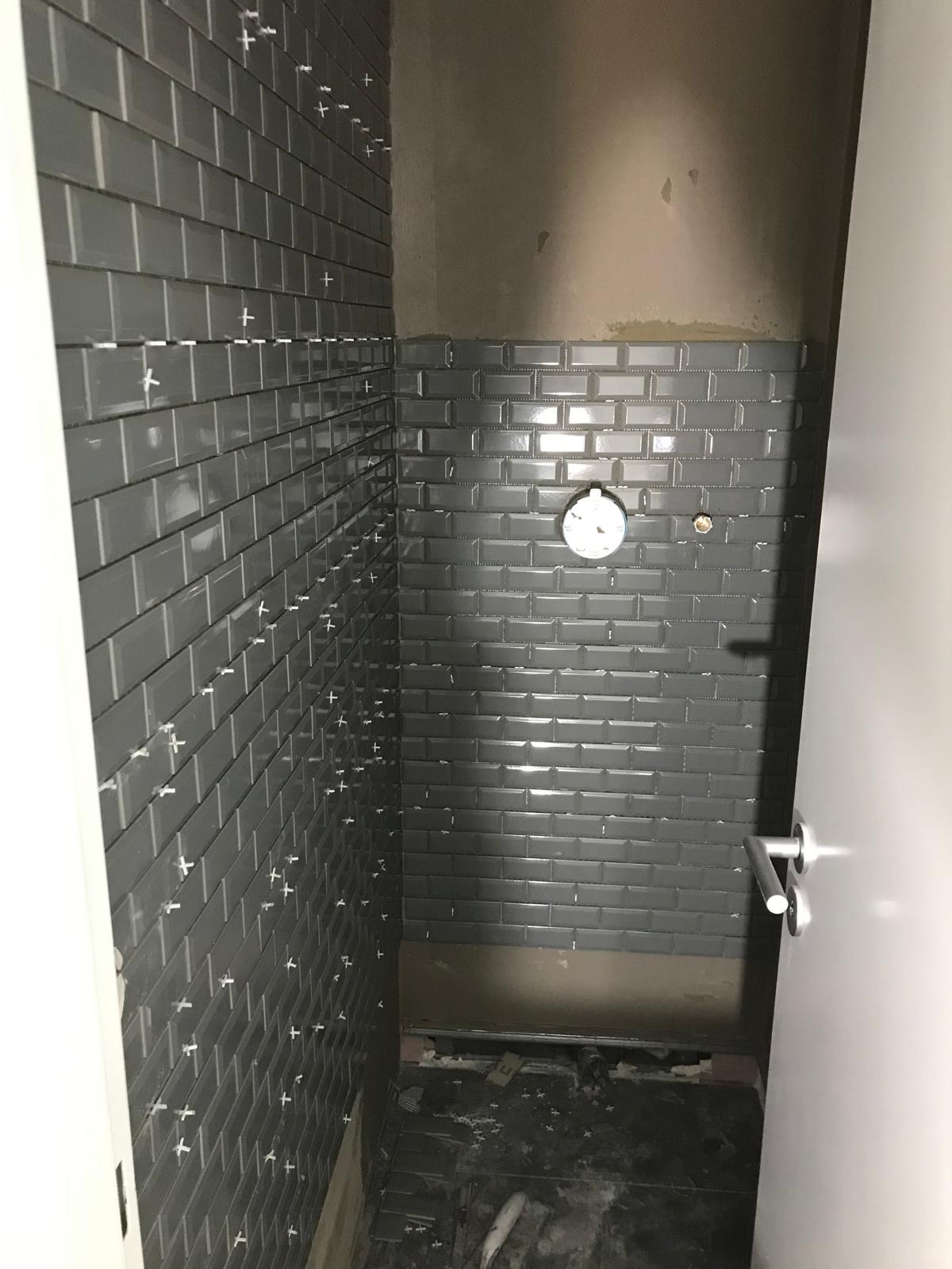 Ściana łazienki w trakcie układania szarych, błyszczących płytek typu metro z białymi krzyżykami dystansowymi. Częściowo wykończona ściana i surowy stan podłogi.