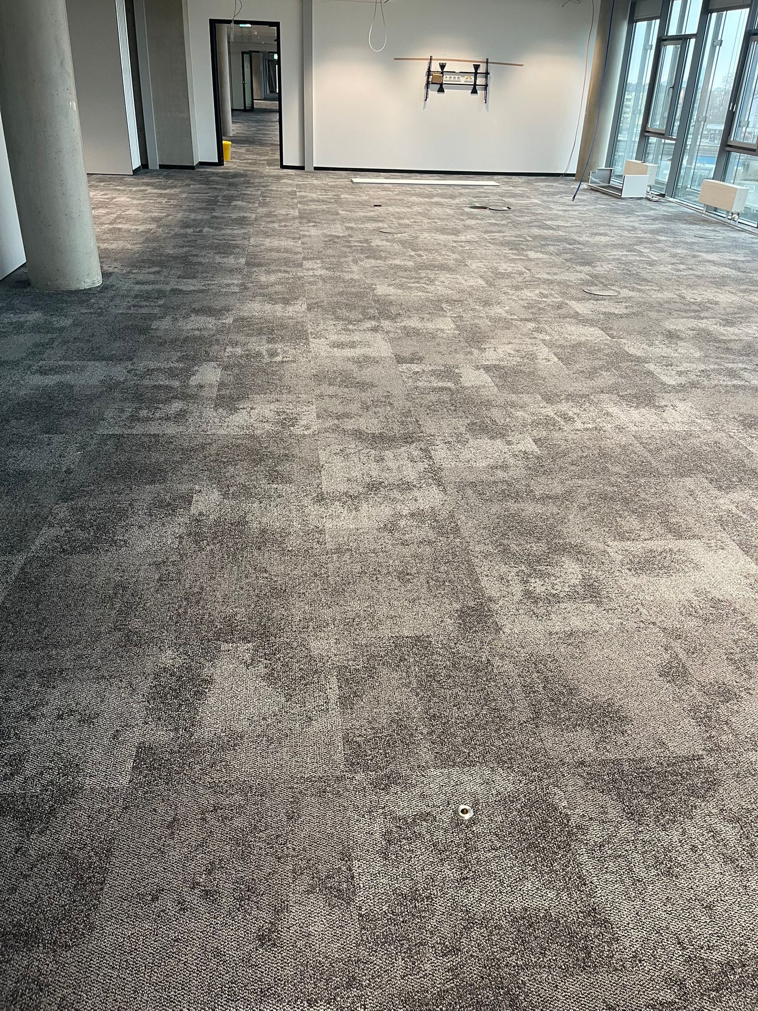 Szara wykładzina dywanowa w biurze z kolumną i oknami. Widok na całą powierzchnię, wzór w kwadraty, jasne ściany, surowy stan wykończenia.