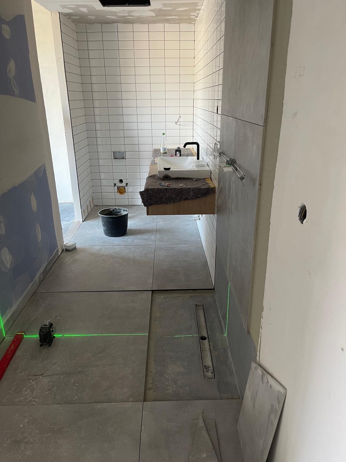 Łazienka w trakcie prac: ułożone płytki podłogowe, częściowo wykończone ściany z białymi kaflami, umywalka z czarną armaturą. Widoczny laser krzyżowy.