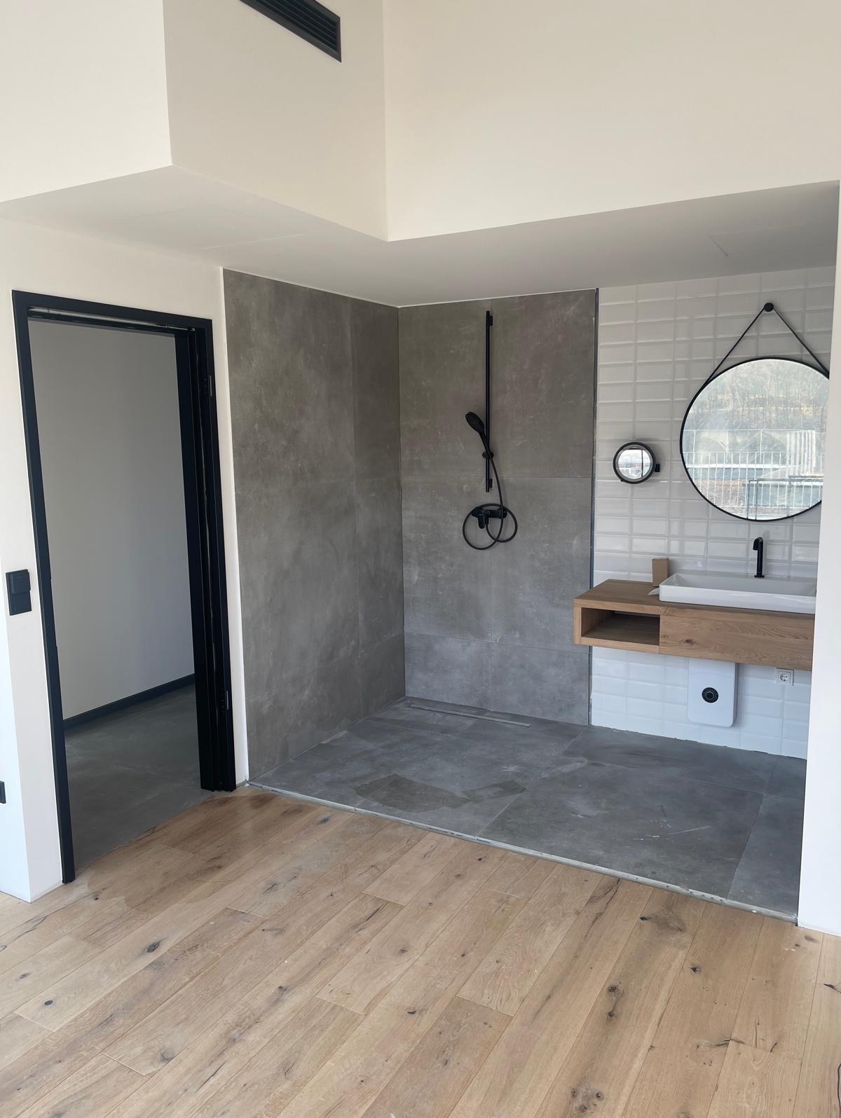 Nowoczesna łazienka z szarymi płytkami, drewnianą szafką pod umywalką i okrągłym lustrem. Podłoga wyłożona drewnianym parkietem. Minimalistyczny design.