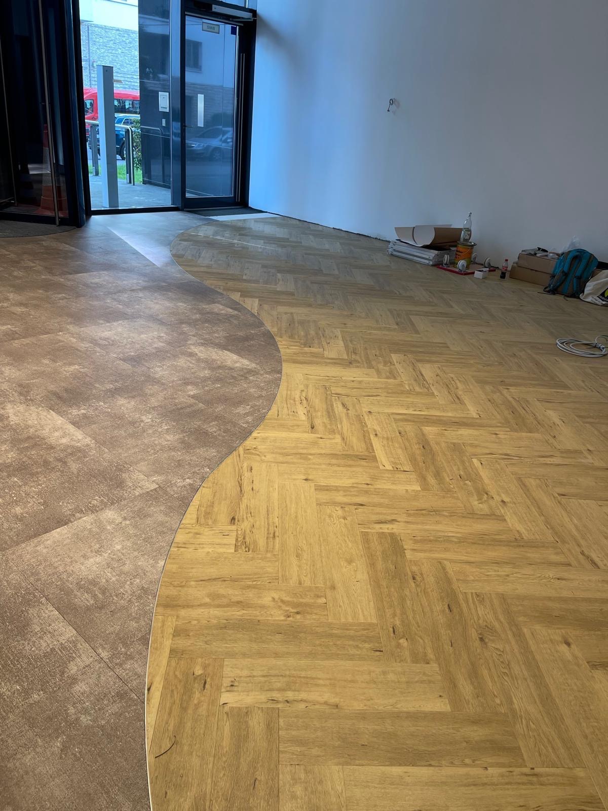 Ukończona podłoga z dwoma rodzajami paneli, ułożona w jodełkę i z zaokrąglonym przejściem. Widoczne materiały i narzędzia w tle, przy białej ścianie.