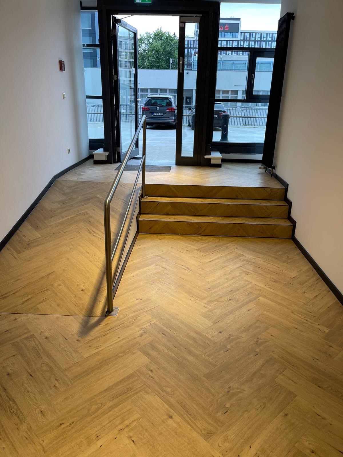 Nowa podłoga z parkietu ułożonego w jodełkę w korytarzu z trzema schodami i metalową barierką. Widok na zewnątrz przez otwarte drzwi.