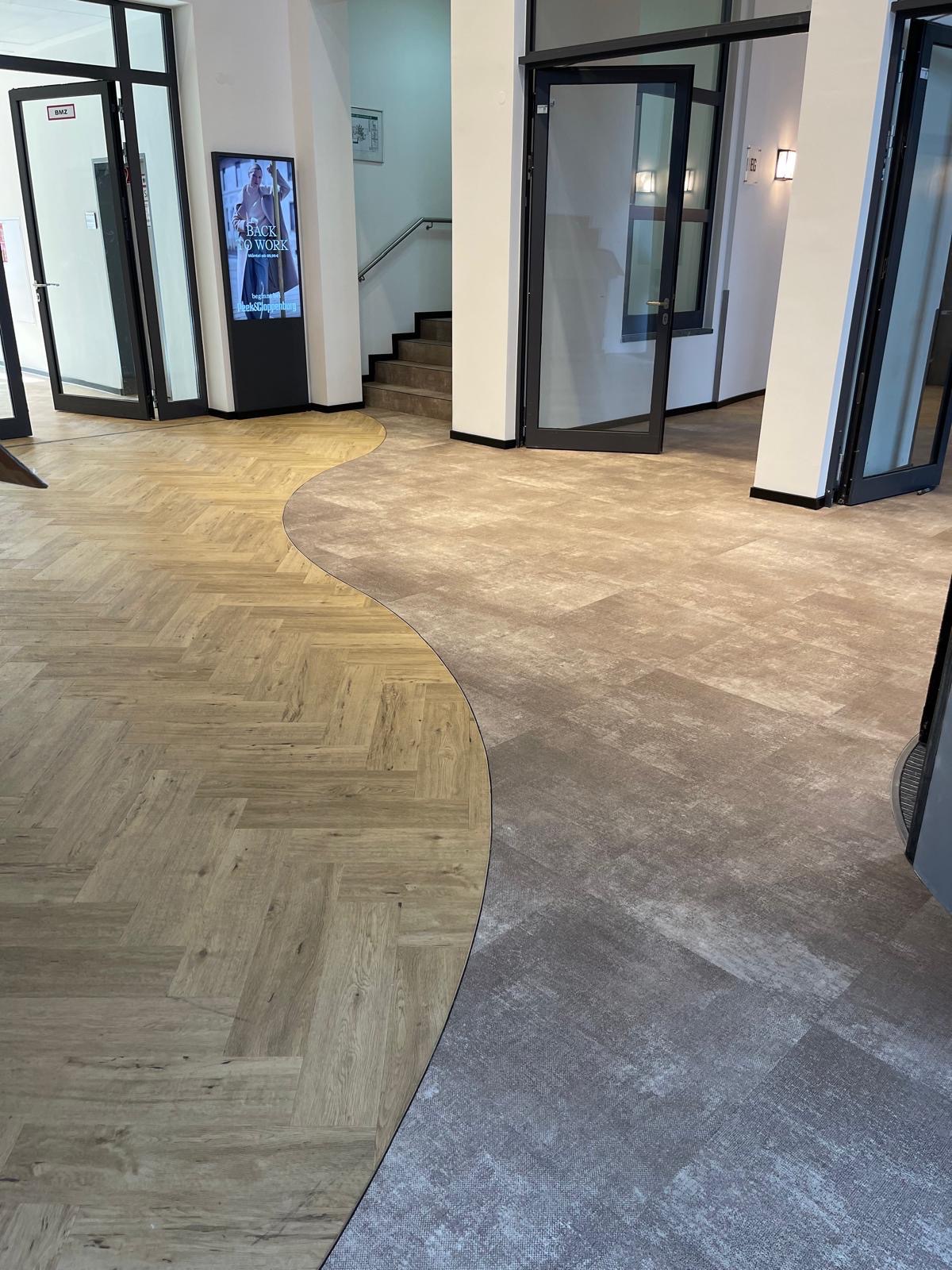 Nowoczesne wykończenie podłogi w biurze z połączeniem parkietu w jodełkę i płytek imitujących beton, oddzielonych falistą linią. Widoczne drzwi i schody w tle.