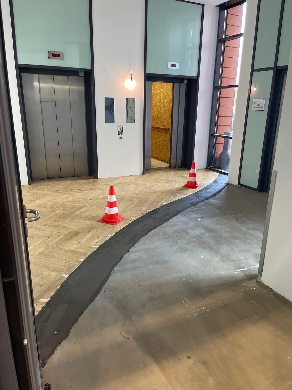 Hol w budynku w trakcie prac wykończeniowych: ułożona podłoga z płytek imitujących drewno, wylewka oddzielona taśmą i pachołkami. Widoczne windy i okna.