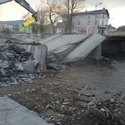 TERRADEM - Rozbiórka mostu nad rzeką z użyciem młota hydraulicznego Volvo. Gruz i fragmenty betonu w korycie rzeki. W tle budynki i drzewa.