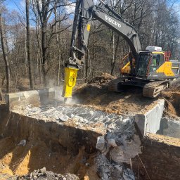TERRADEM - Żółta koparka Volvo z młotem pneumatycznym kruszy betonowy fundament na tle drzew. Widoczne zbrojenia i gruz.