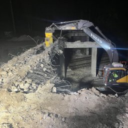 TERRADEM - Żółto-szara koparka Volvo z młotem hydraulicznym kruszy betonowy wiadukt kolejowy nocą. Widoczne zbrojenia i gruz. Operator w kabinie.
