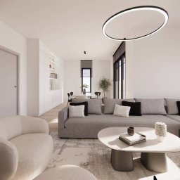 PROJEKTOWANIE WNĘTRZ Adrianna Lis - Jasne wnętrze z szarym narożnikiem, okrągłym stolikiem kawowym i designerską lampą. Minimalistyczny styl, neutralna kolorystyka, widok na jadalnię w tle.