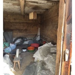Przeprowadzki - Zabrudzone, drewniane pomieszczenie z różnymi przedmiotami, w tym plastikowymi miskami, kablami i folią. Widoczny mały stołek. Nierówna podłoga i ściany.