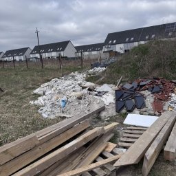 Przeprowadzki - Sterta gruzu, dachówek i drewna na zewnątrz, z rzędem domów w tle pod pochmurnym niebem. Widoczne ślady po demontażu, teren przed uprzątnięciem.