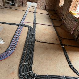 elektrykTAKt - Kable elektryczne w peszlach ułożone na podłodze w surowym wnętrzu z ceglanymi ścianami. Widoczne mocowania kabli. Etap prac instalacyjnych przed wykończeniem.
