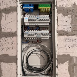 elektrykTAKt - Otwarta skrzynka elektryczna z przewodami i bezpiecznikami, zamontowana na surowej ścianie z widocznymi napisami. Widoczne elementy instalacji elektrycznej.