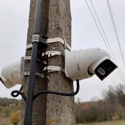elektrykTAKt - Dwie kamery Dahua zamontowane na słupie w Warszawie. Kamery przymocowane metalowymi obejmami, widoczne przewody. Tło stanowią drzewa i pochmurne niebo.