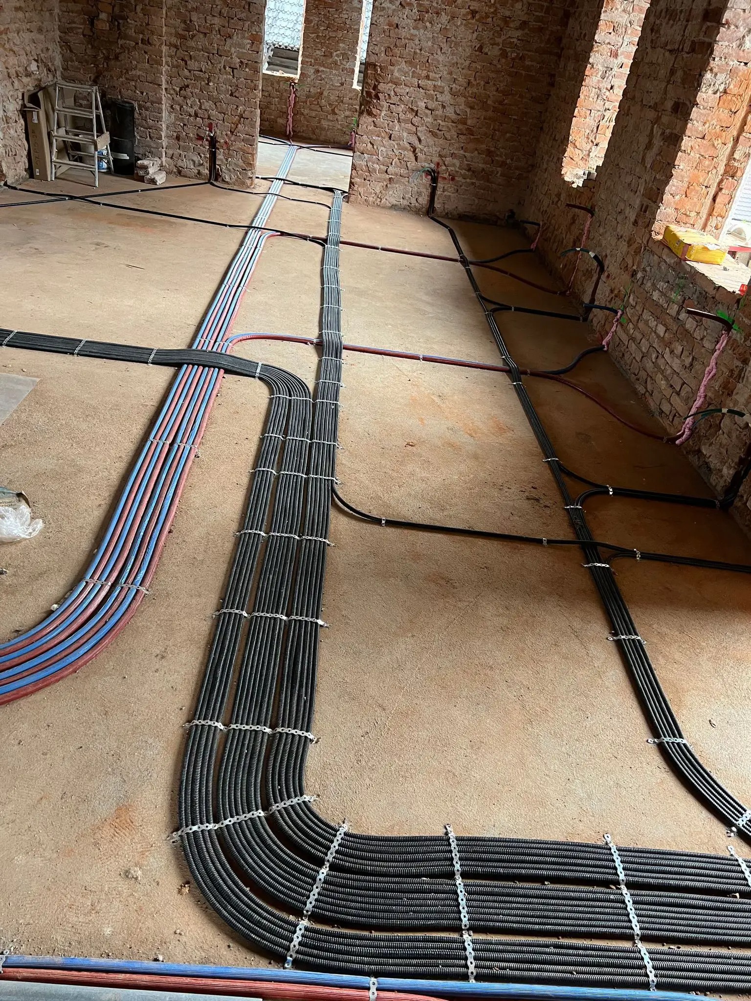 Kable elektryczne w peszlach ułożone na podłodze w surowym wnętrzu z ceglanymi ścianami. Widoczne mocowania kabli. Etap prac instalacyjnych przed wykończeniem.