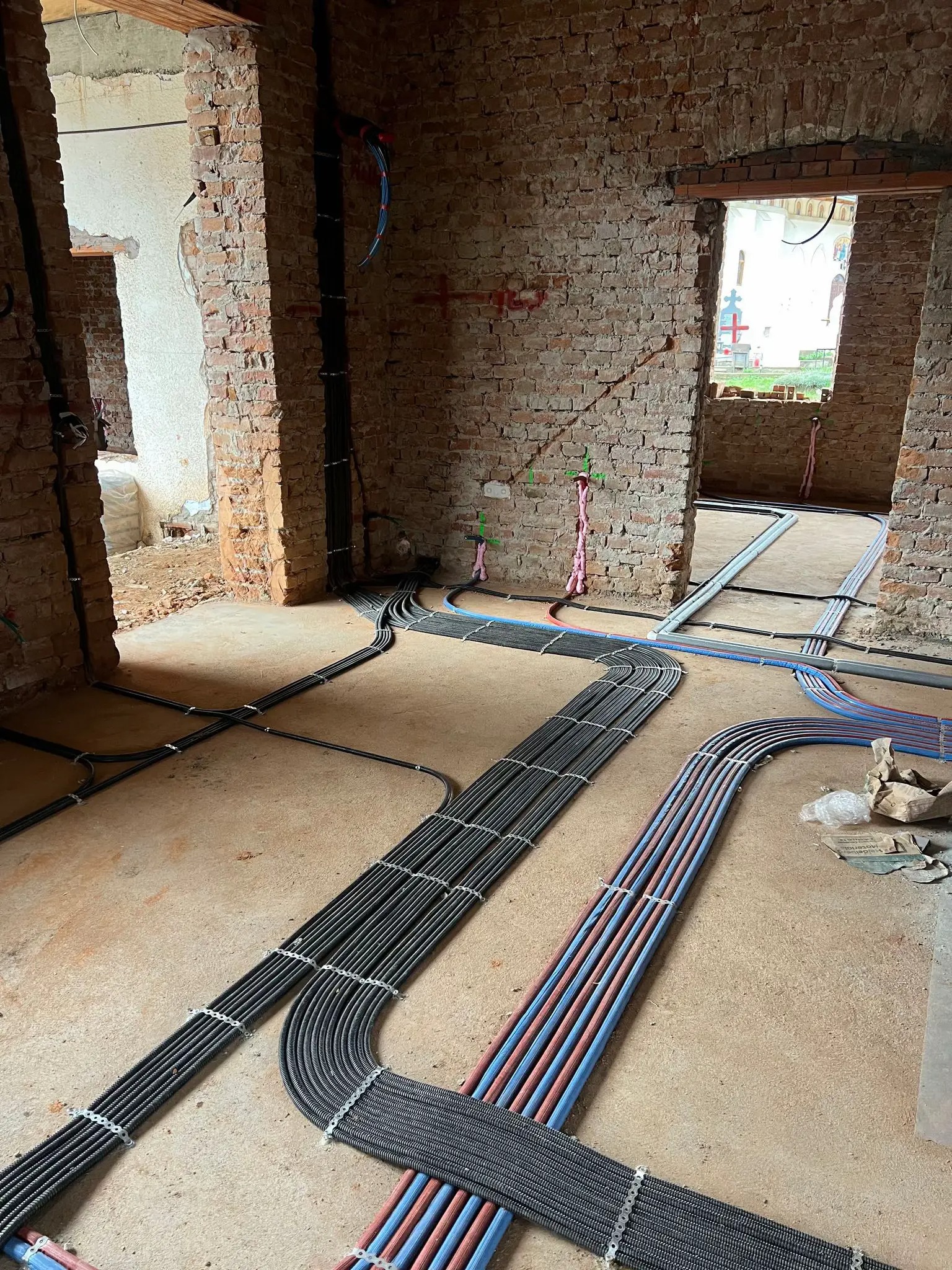 Wnętrze w trakcie remontu: ułożone rury instalacyjne (czerwone, niebieskie, czarne) na podłodze, ściany z cegły, widoczne otwory drzwiowe. Etap prac instalacyjnych.