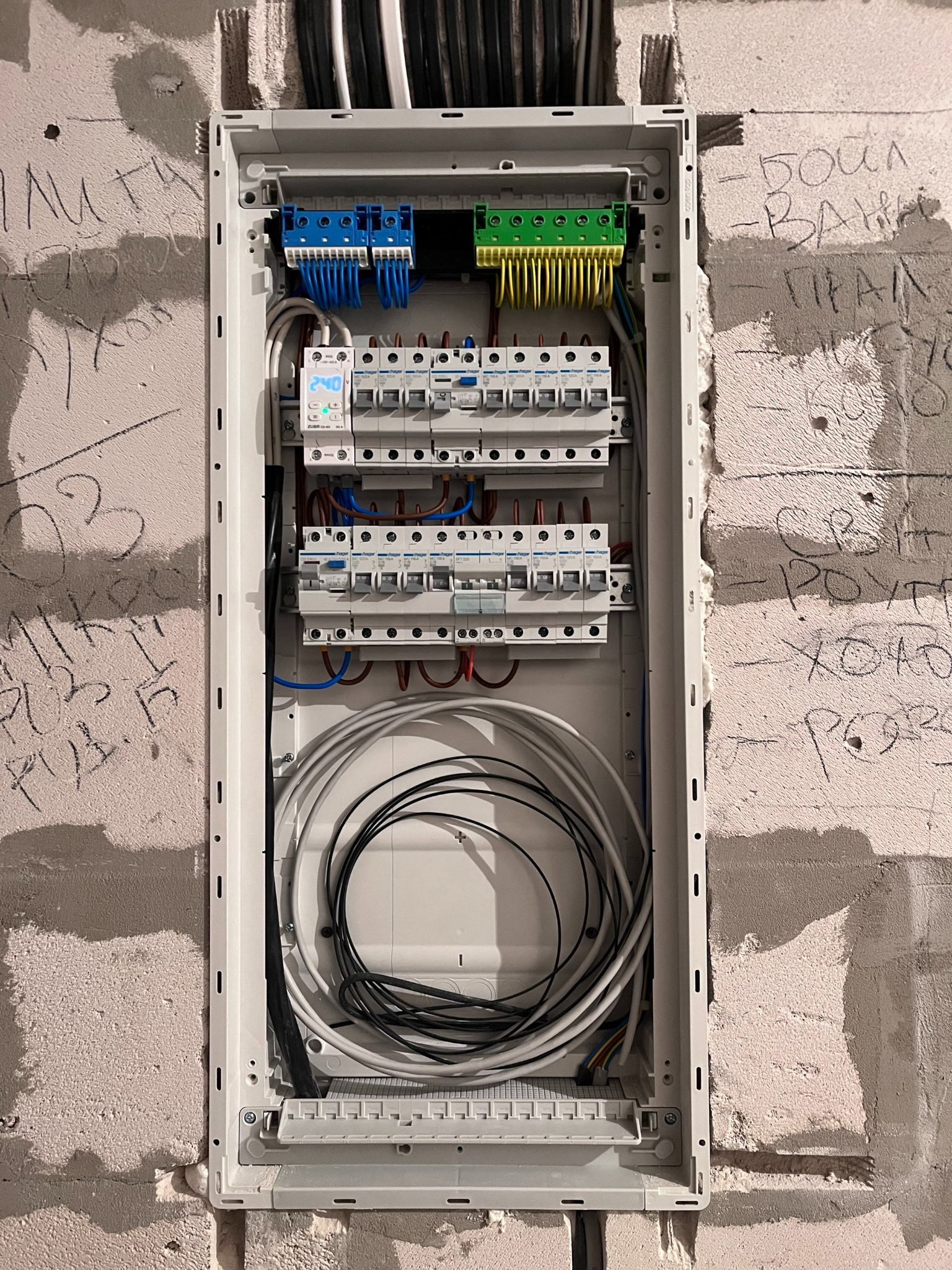 Otwarta skrzynka elektryczna z przewodami i bezpiecznikami, zamontowana na surowej ścianie z widocznymi napisami. Widoczne elementy instalacji elektrycznej.