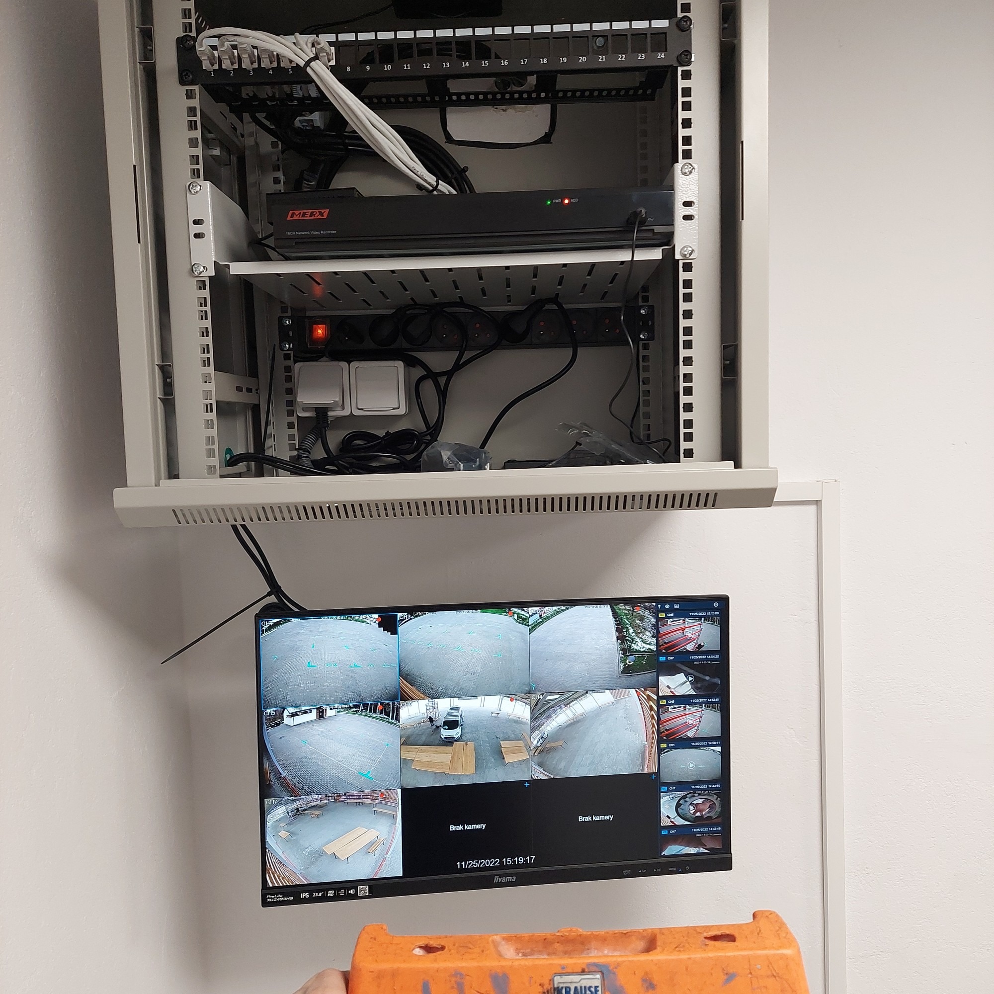 Szafa rack z rejestratorem MERX i patch panelem, poniżej listwa zasilająca i monitor z podglądem z kamer monitoringu. Widoczne kable i pomarańczowa barierka.