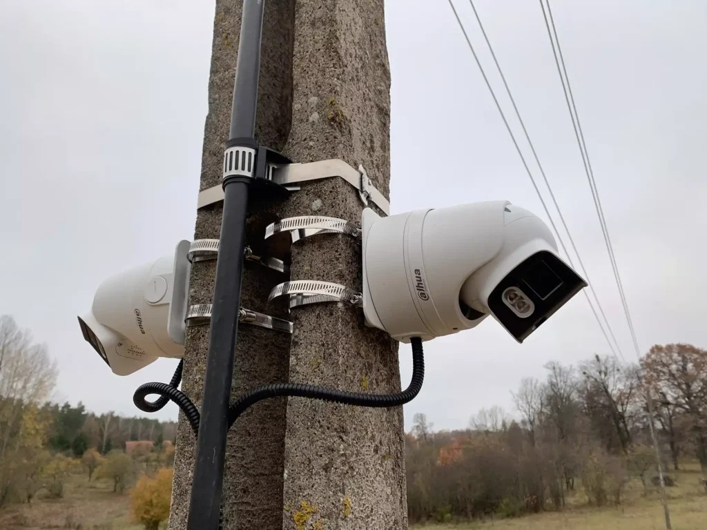 Dwie kamery Dahua zamontowane na słupie w Warszawie. Kamery przymocowane metalowymi obejmami, widoczne przewody. Tło stanowią drzewa i pochmurne niebo.