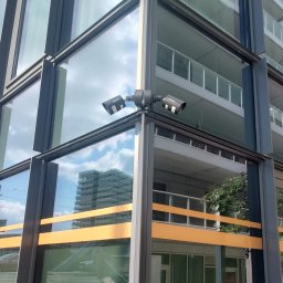SKSYSTEMS SPÓŁKA Z OGRANICZONĄ ODPOWIEDZIALNOŚCIĄ - Dwie kamery monitoringu Hikvision zamontowane na rogu nowoczesnego budynku z przeszkloną fasadą i balkonami. W tle widoczne odbicie nieba i inny budynek.
