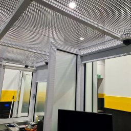 SKSYSTEMS SPÓŁKA Z OGRANICZONĄ ODPOWIEDZIALNOŚCIĄ - Białe kamery monitoringu kopułkowe zamontowane pod ażurowym sufitem w biurze. Widoczne okna i elementy wyposażenia. Żółte pasy na ścianach.