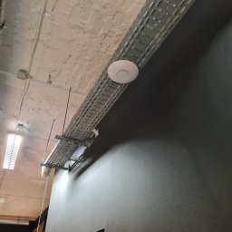 SKSYSTEMS SPÓŁKA Z OGRANICZONĄ ODPOWIEDZIALNOŚCIĄ - Montaż punktu dostępowego WiFi na metalowej trasie kablowej pod sufitem. Widoczne rury wentylacyjne i oświetlenie. Szara ściana w tle.