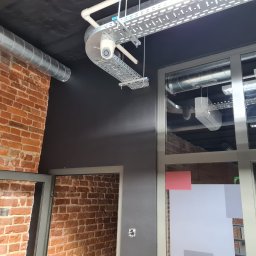 SKSYSTEMS SPÓŁKA Z OGRANICZONĄ ODPOWIEDZIALNOŚCIĄ - Kamera monitoringu zamontowana na metalowej konstrukcji pod sufitem, widoczna instalacja wentylacyjna i fragment ściany z czerwonej cegły w industrialnym wnętrzu.