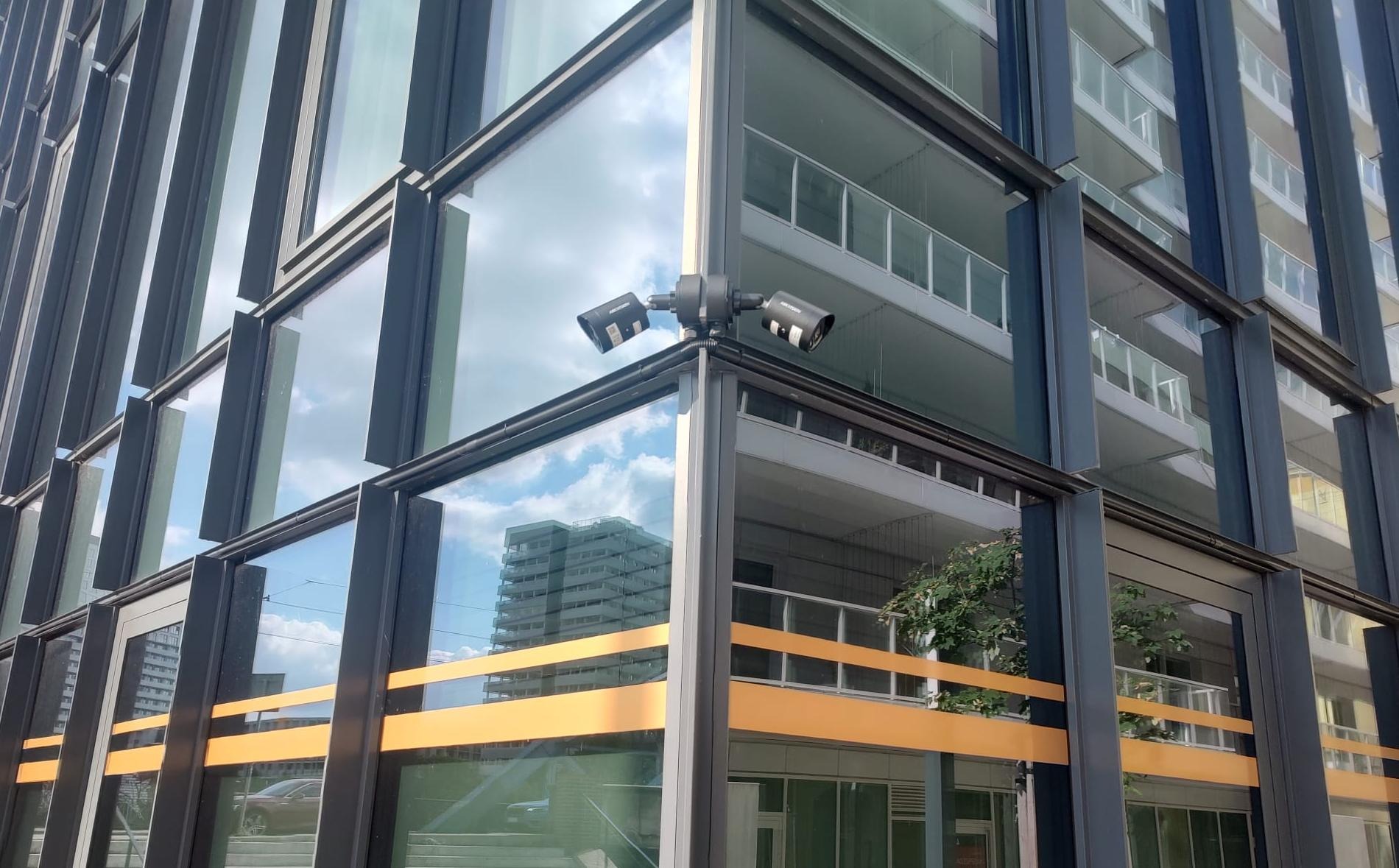Dwie kamery monitoringu Hikvision zamontowane na rogu nowoczesnego budynku z przeszkloną fasadą i balkonami. W tle widoczne odbicie nieba i inny budynek.