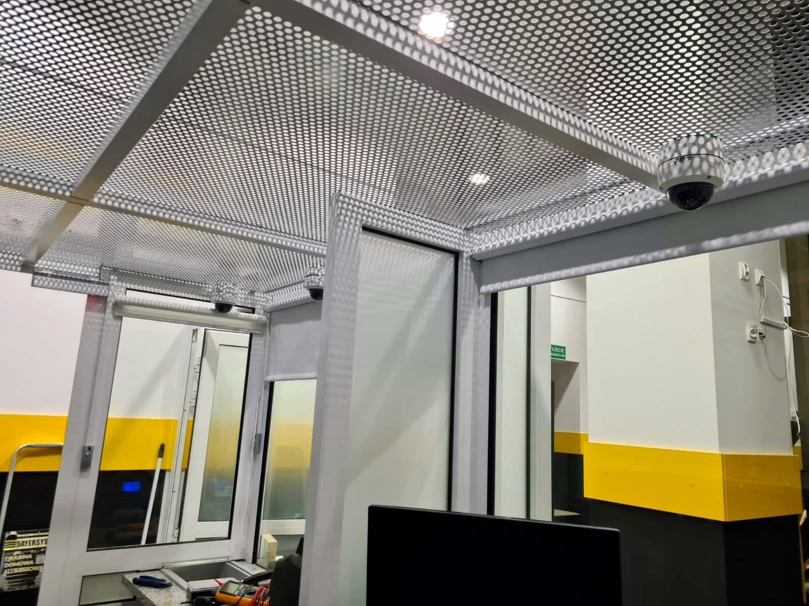 Białe kamery monitoringu kopułkowe zamontowane pod ażurowym sufitem w biurze. Widoczne okna i elementy wyposażenia. Żółte pasy na ścianach.
