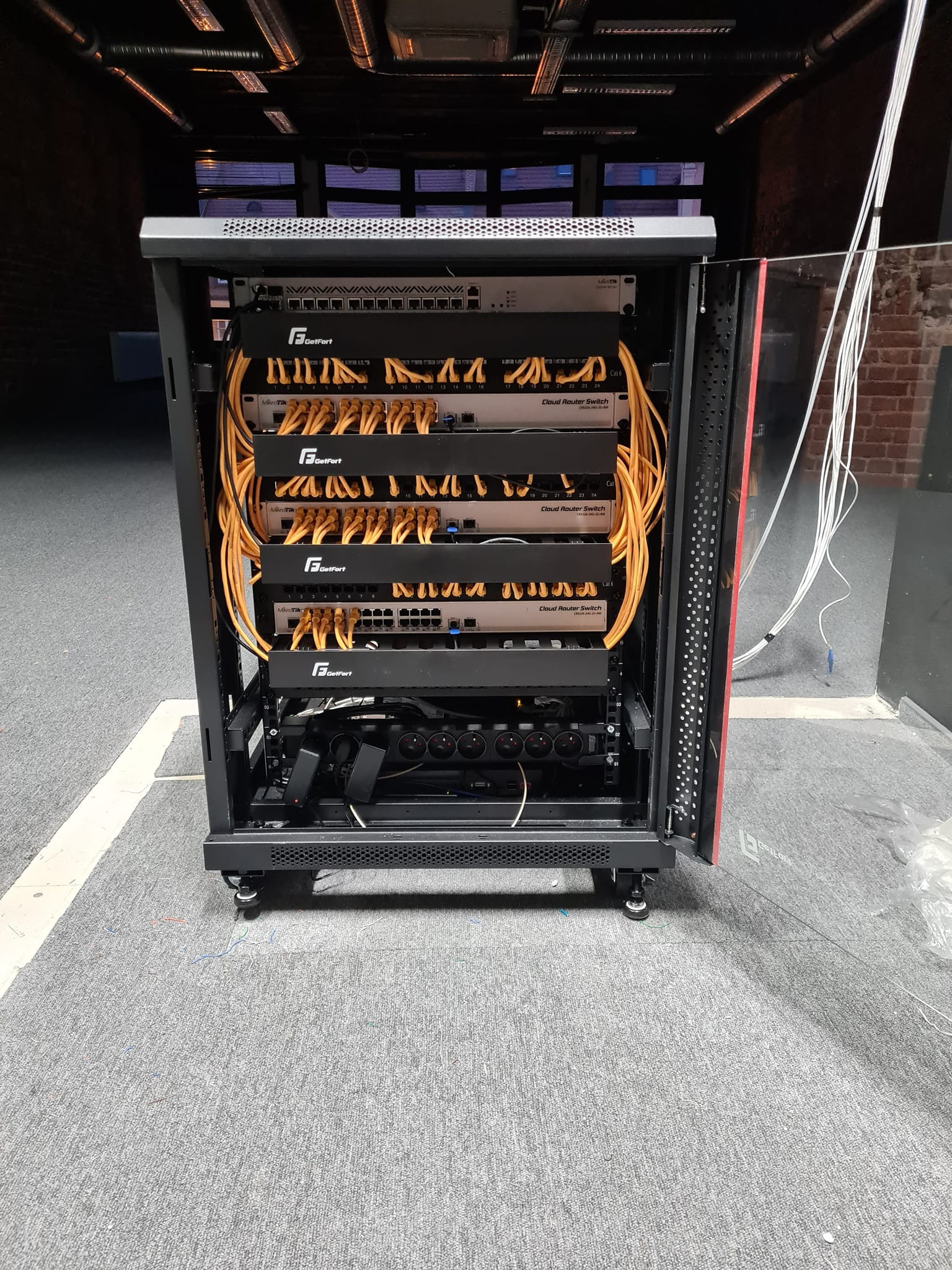 Otwarta szafa rack z zamontowanymi routerami, switchami i okablowaniem. Widoczne urządzenia marki GetFort i MikroTik. Kable patchcord w kolorze żółtym.