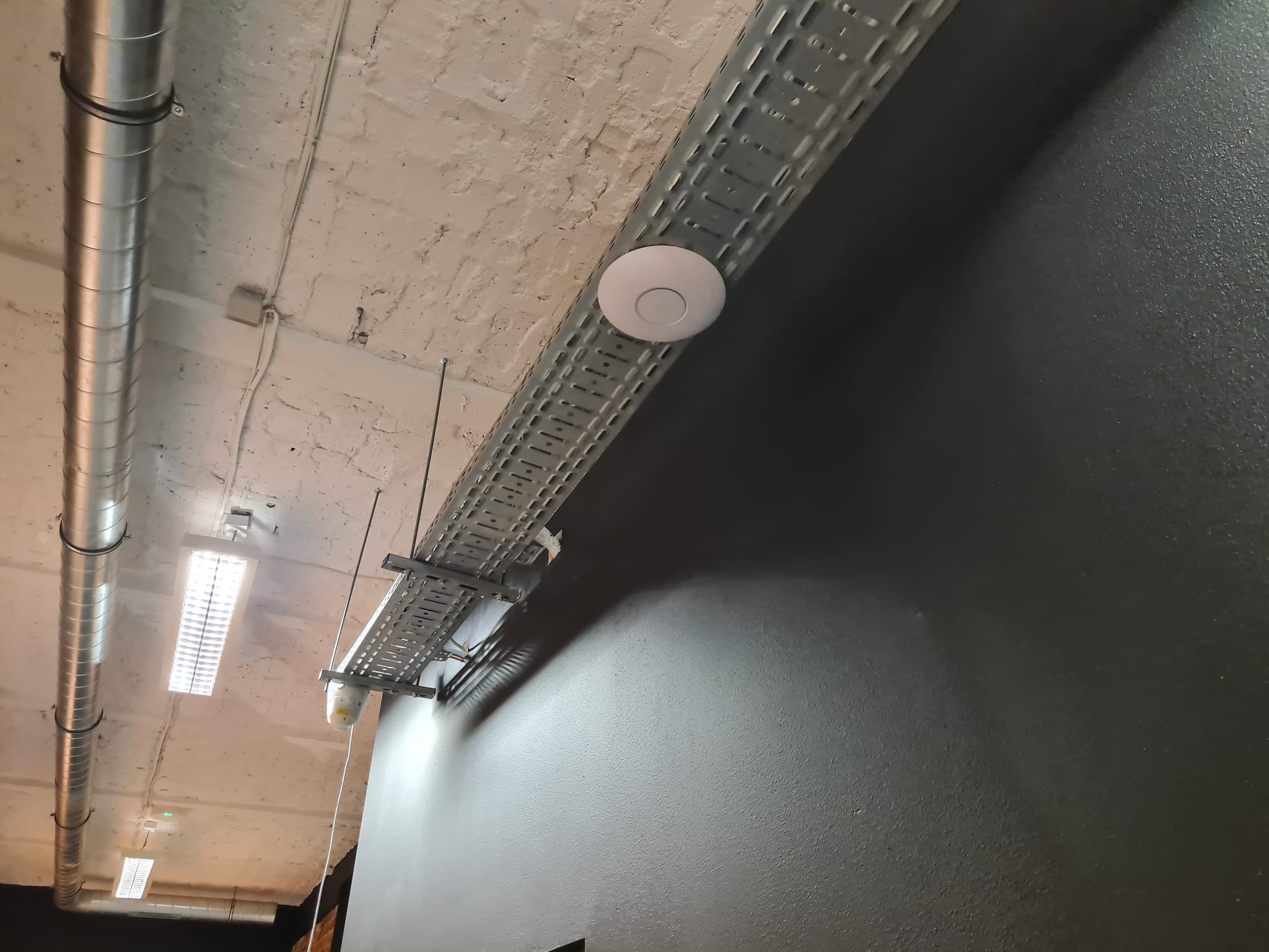 Montaż punktu dostępowego WiFi na metalowej trasie kablowej pod sufitem. Widoczne rury wentylacyjne i oświetlenie. Szara ściana w tle.