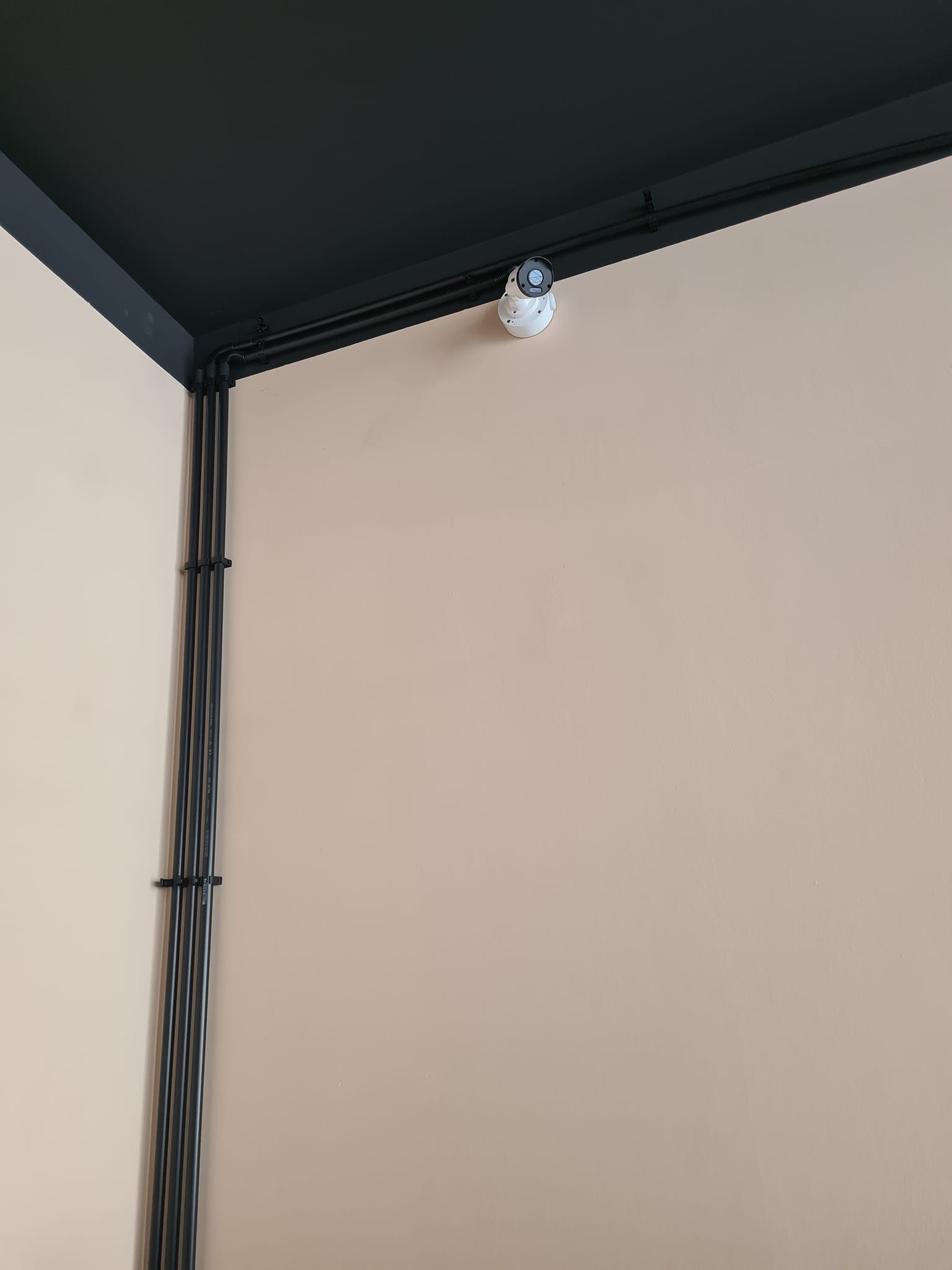 Kamera monitoringu zamontowana w rogu pomieszczenia z czarnym sufitem i beżowymi ścianami. Widoczne czarne rurki instalacyjne przy ścianie.