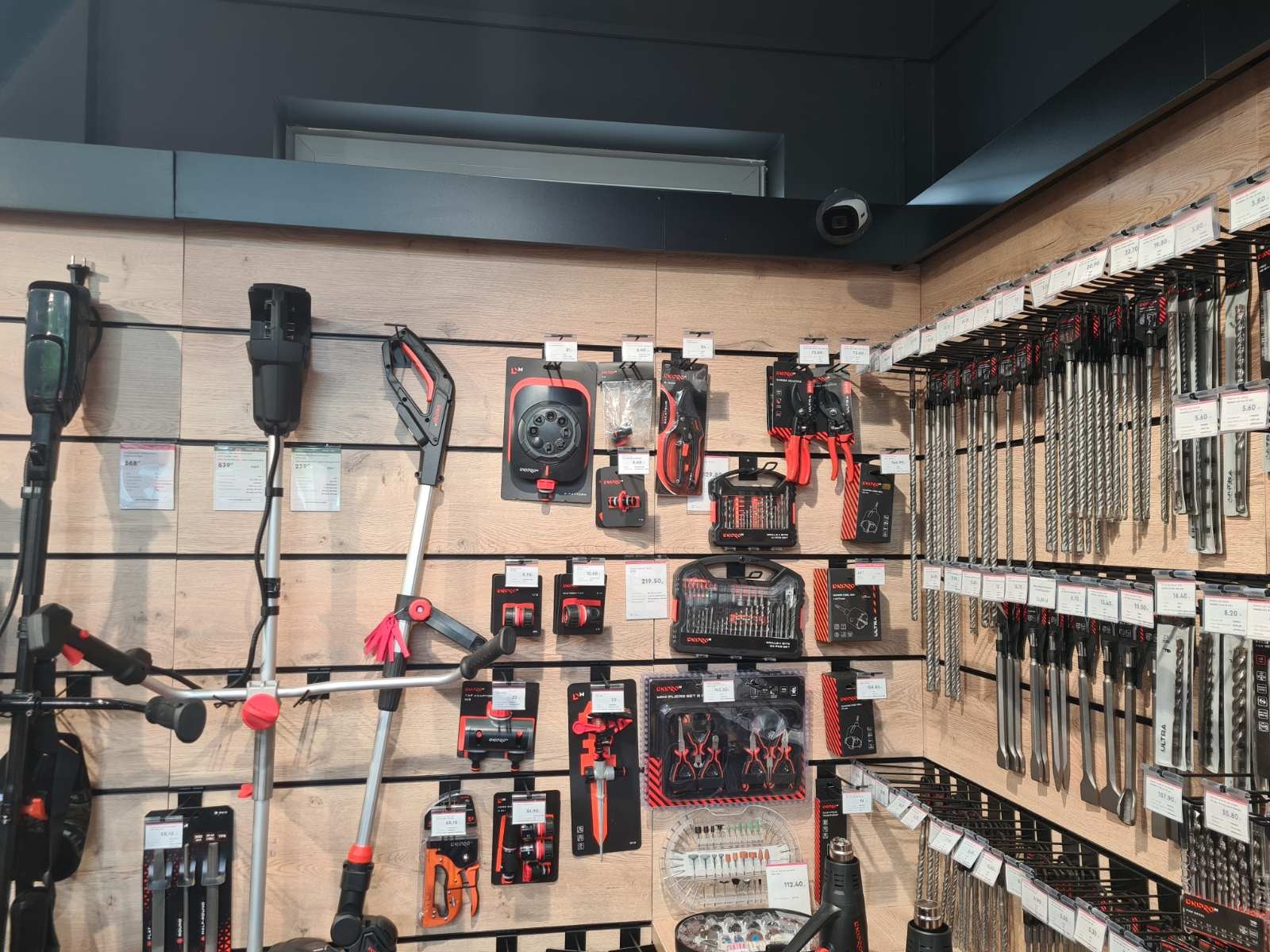 Ekspozycja narzędzi ogrodowych i wierteł w sklepie: kosiarki, zestawy narzędzi, wiertła różnych rozmiarów. Widoczna kamera monitoringu pod sufitem.