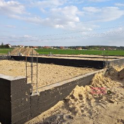 DMP - Fundamenty domu jednorodzinnego z izolacją przeciwwilgociową, częściowo zasypane piaskiem, z wystającymi prętami zbrojeniowymi. W tle zielone pola i zabudowa wiejska.