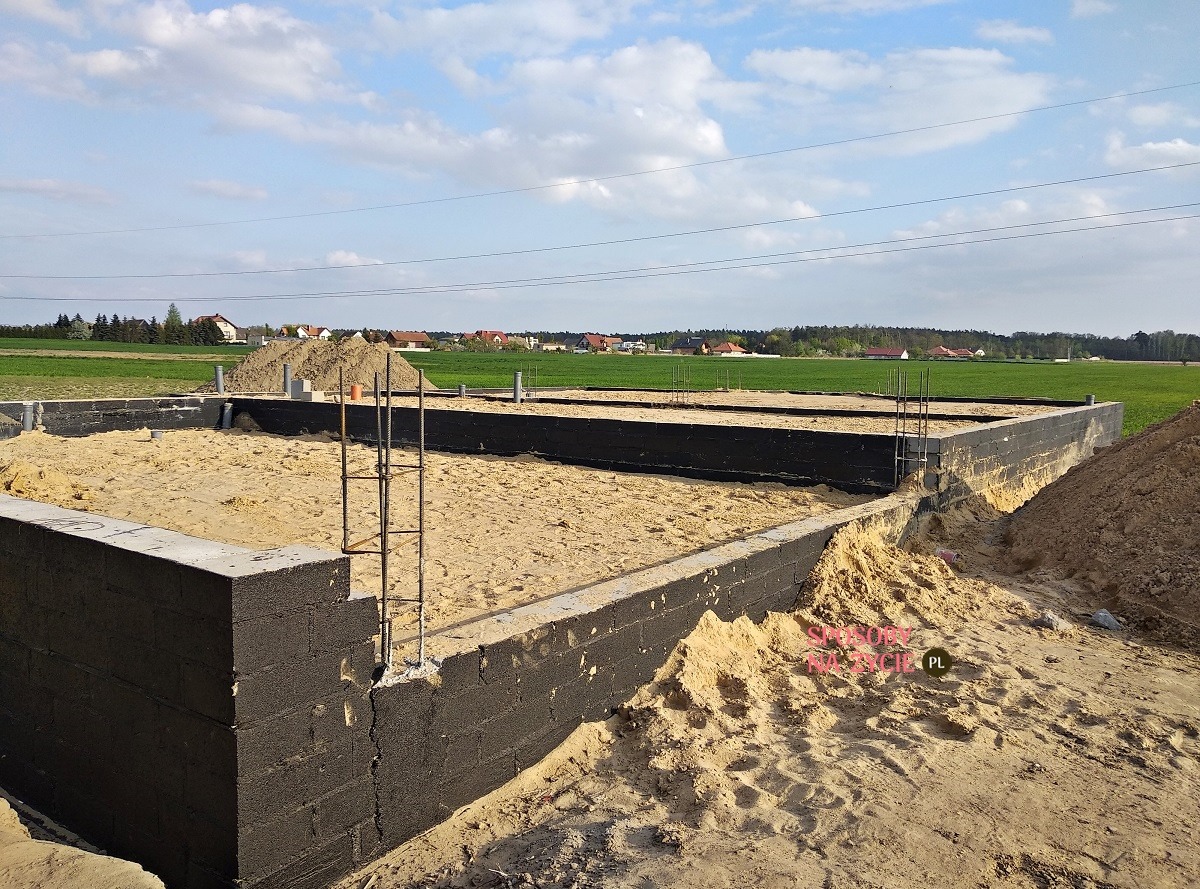 Fundamenty domu jednorodzinnego z izolacją przeciwwilgociową, częściowo zasypane piaskiem, z wystającymi prętami zbrojeniowymi. W tle zielone pola i zabudowa wiejska.